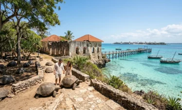 A dynamic horizontal documentary photograph capturing the unique historical and natural environment of Prison Island (Changuu), off the coast of Stone Town, Zanzibar, during a bright afternoon. It features massive Aldabra Giant Tortoises, weathered coral-rag limestone ruins with arched windows and tile roofs, a long wooden pier over shallow crystal-clear turquoise water, anchored traditional dhow boats, dense coastal vegetation, and the distant Stone Town skyline. Natural daylight filters through trees.