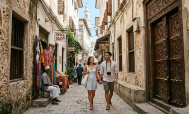 A high-resolution candid documentary photograph of a narrow, sun-drenched alleyway in Stone Town, Zanzibar, featuring traditional coral-rag limestone buildings, an intricate hand-carved teak Zanzibar door, and a local resident walking past a small spice shop, capturing the authentic UNESCO World Heritage atmosphere.